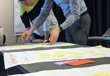 Two people leaning over flip chart pages on a table, interacting with post-it notes.