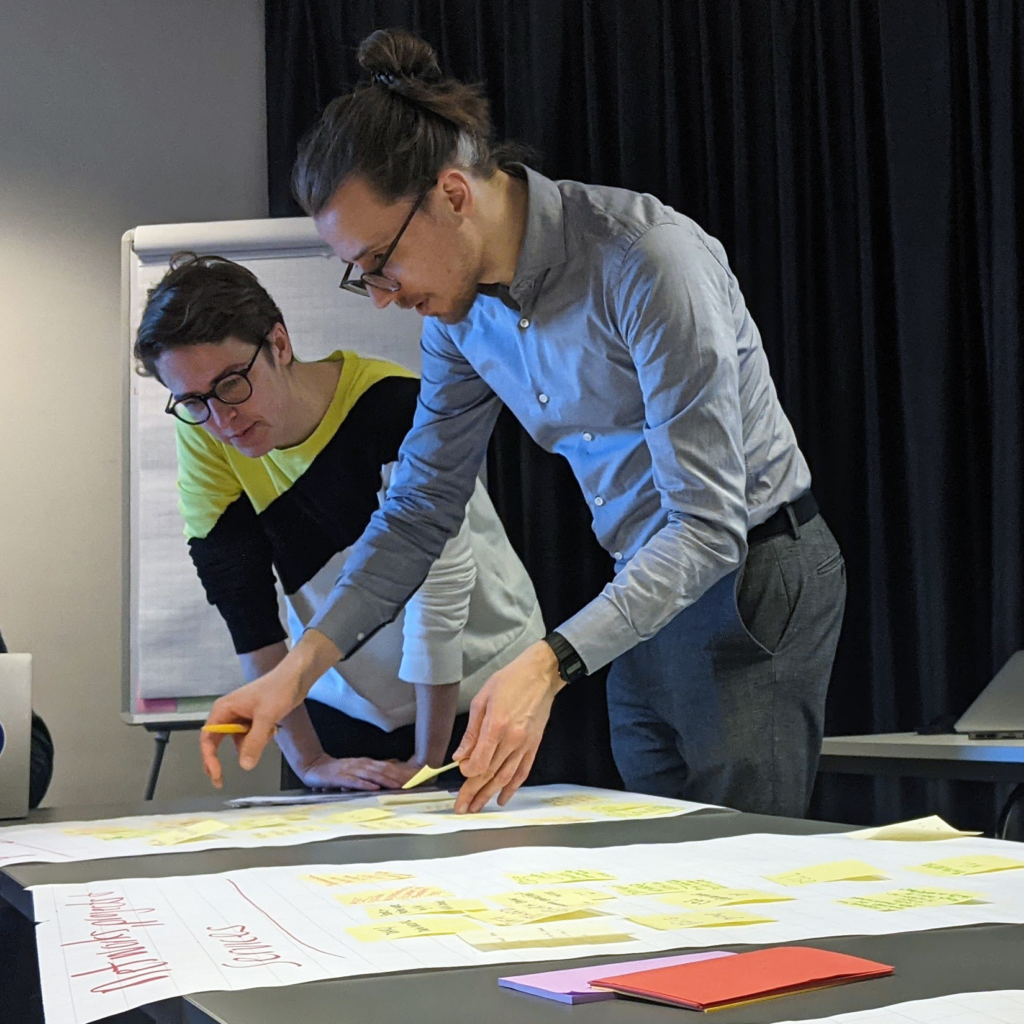 Two people leaning over flip chart pages on a table, interacting with post-it notes.