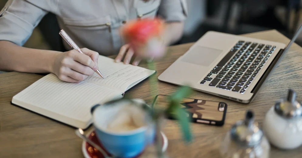 A person sitting at a desk in front of a laptop, taking notes on a notepad.